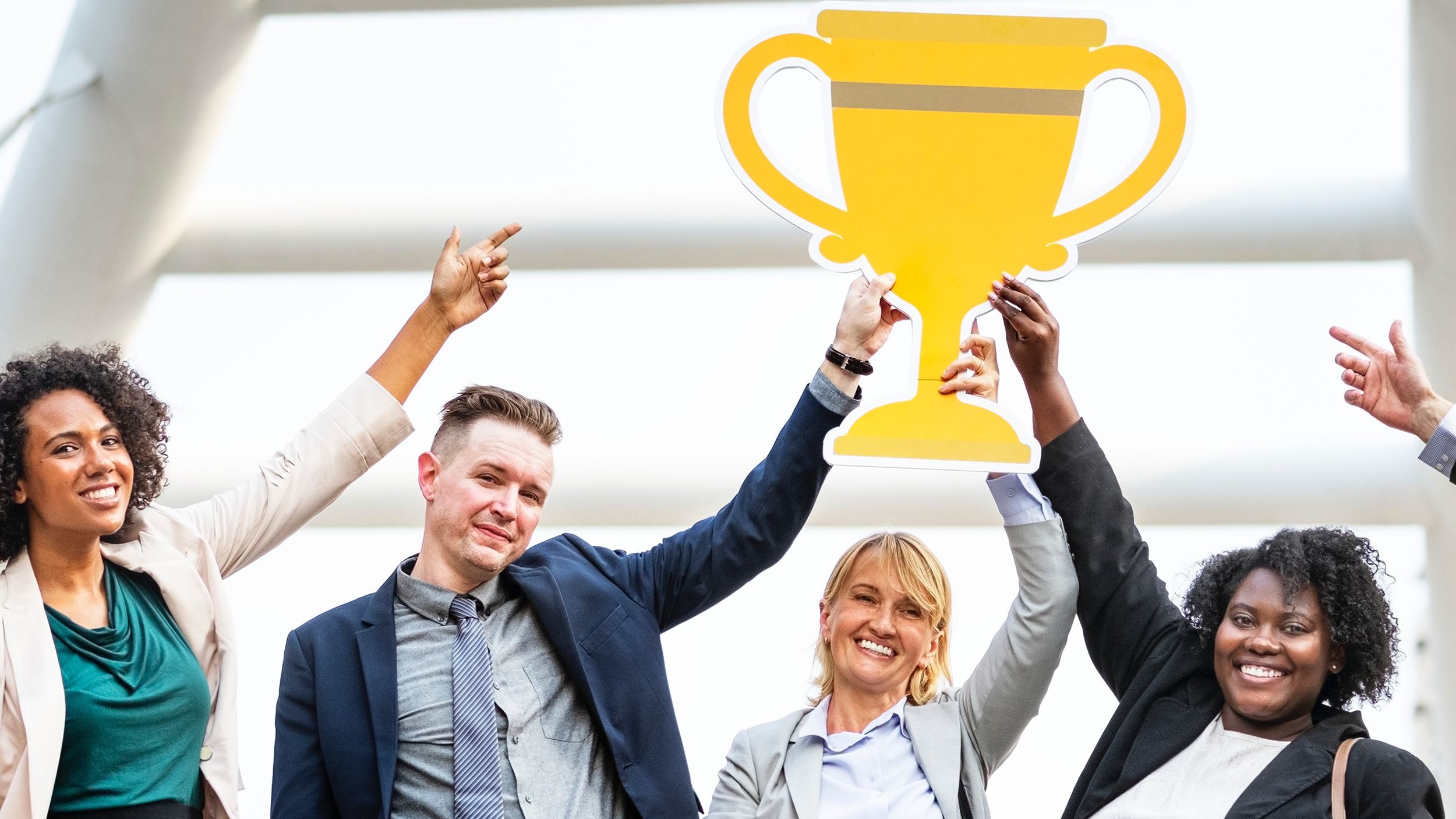 Four diverse professionals smiling and celebrating while holding a large yellow trophy cutout together, representing teamwork and success